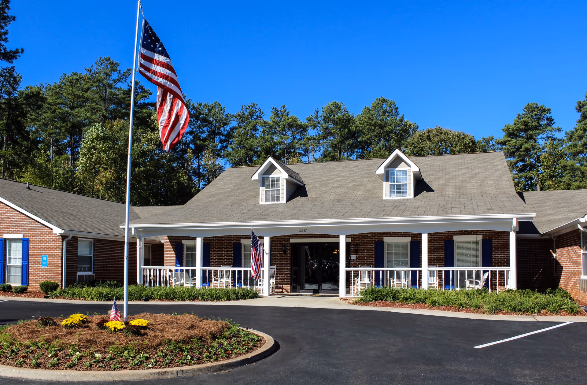 Front exterior view of a single-story brick building with a gray shingled roof and two dormer windows. The building has a covered porch with white railings and several white rocking chairs. An American flag is flying on a flagpole in front of a circular flower bed with yellow flowers. Trees and a clear blue sky are visible in the background.