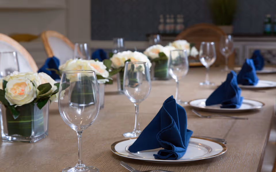 A dining table set with white plates, blue folded napkins, empty wine glasses, and floral centerpieces with white roses in glass vases. The background shows chairs and a sideboard with decorative items.