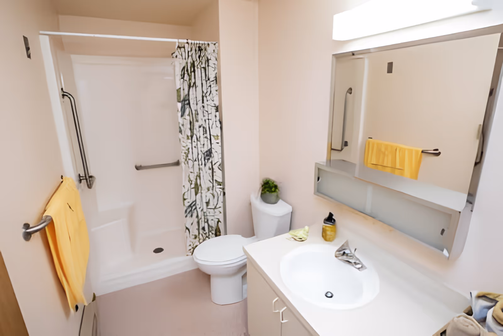 A clean bathroom with a white toilet, a white sink with a silver faucet, a large mirror above the sink, and a shower area with a patterned shower curtain. There is a yellow towel hanging on a towel rack next to the shower and a small green plant on top of the toilet tank.