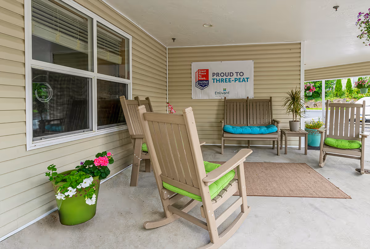 Covered outdoor seating area with beige rocking chairs and a bench, all with colorful cushions. There are potted plants with flowers placed around the space, and a sign on the wall that reads 'Proud to Three-Peat' with the Enlivant logo. The area has a concrete floor and beige siding walls.