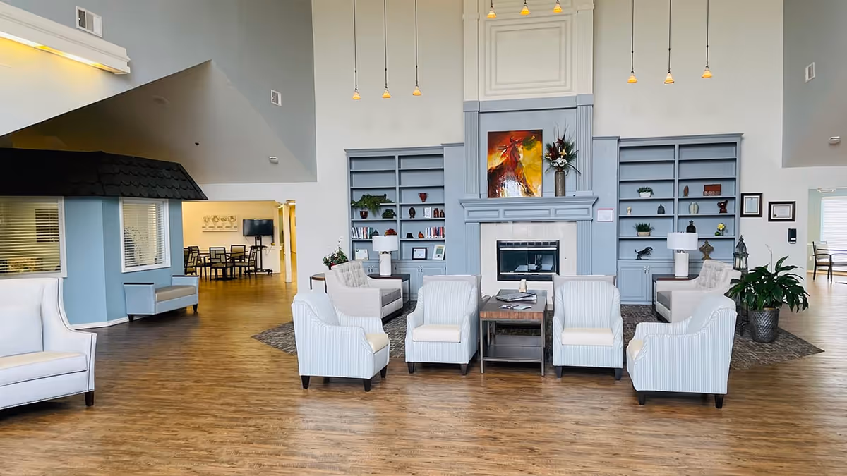 A spacious and well-lit living room area in a senior living facility with light blue walls and built-in shelves flanking a fireplace. The room features several white cushioned armchairs arranged around a wooden coffee table on a rug. There are pendant lights hanging from the high ceiling, a colorful painting above the fireplace, and some plants and decorative items on the shelves. In the background, there is a dining area with tables and chairs.