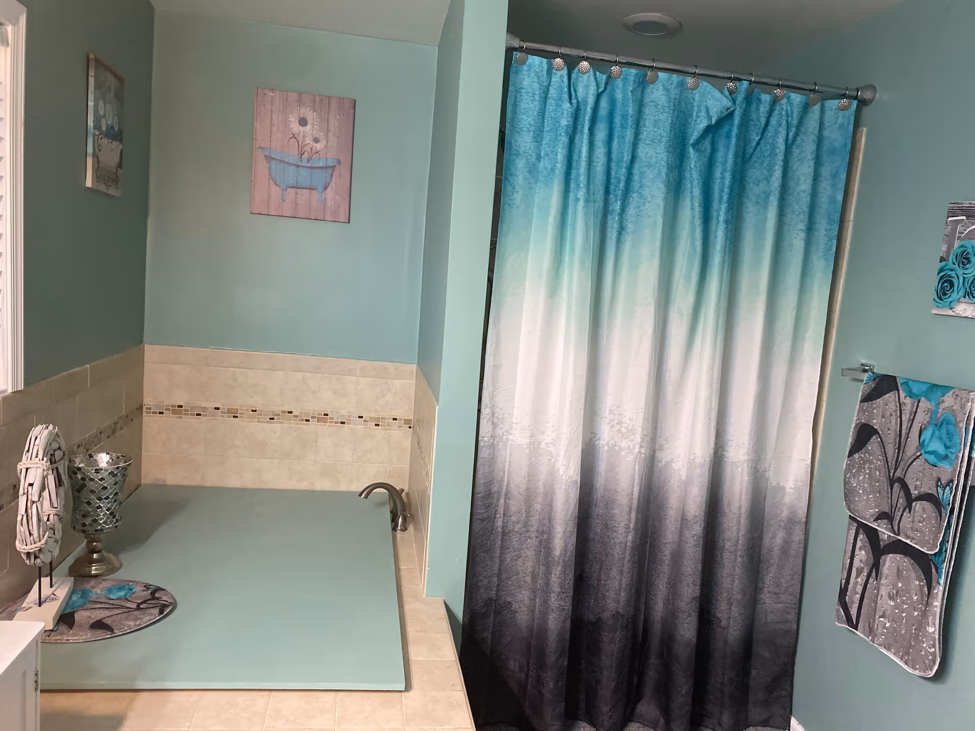 Bathroom with teal walls and beige tiled lower walls. There is a bathtub with a teal-colored bath mat, a silver decorative candle holder, and a small wreath decoration on the edge. A shower curtain with a gradient from teal to white to black hangs on a rod. Two towels with floral patterns in teal, black, and gray hang on a towel rack. Two framed pictures with bath-related themes hang on the walls.
