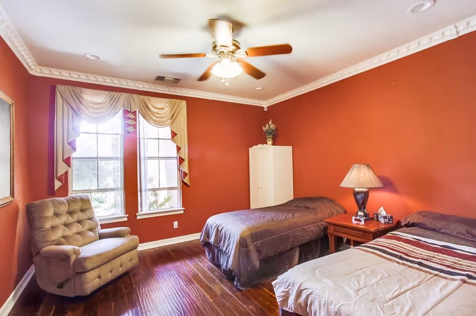 Bright bedroom with two twin beds, a wooden nightstand and lamp, an upholstered chair, ceiling fan, and red walls.