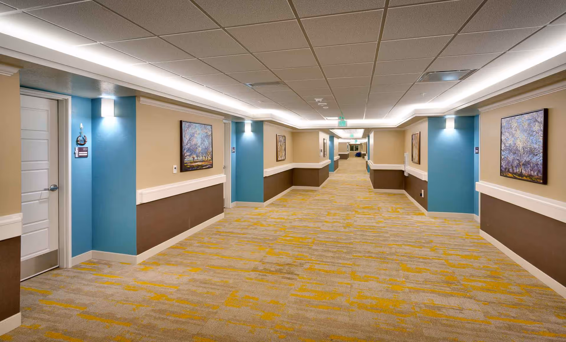 Well-lit interior hallway of a senior living facility with carpeted floors, doors, handrails and framed artwork on the walls.