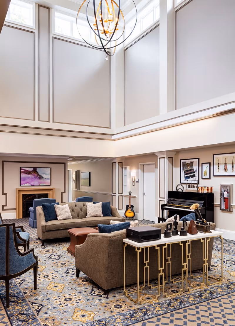 A spacious and elegant living room with high ceilings and large windows near the ceiling. The room features a beige sofa with blue and beige pillows, a matching armchair, a patterned carpet, and a modern chandelier. There is a fireplace with a colorful abstract painting above it, a black upright piano with framed musical instrument pictures on the wall, and a guitar resting against the wall. A console table behind the sofa holds decorative items including a gramophone and a metronome.