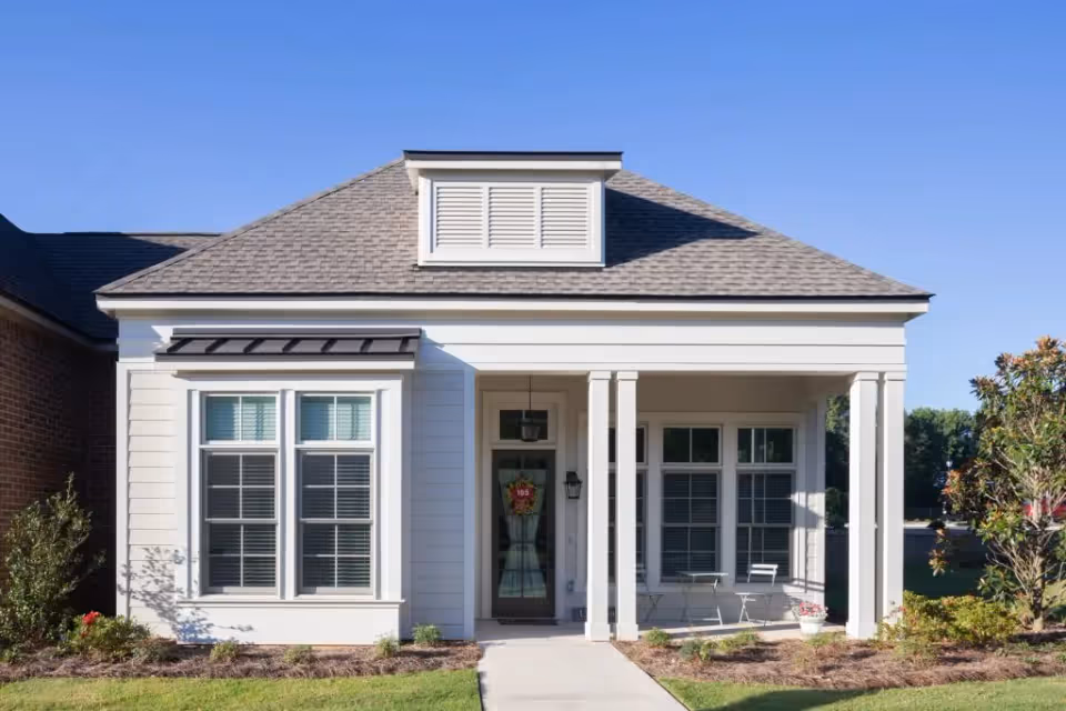 Front exterior view of a single-story residential building with white siding, a gray shingled roof, and a small covered porch with two white columns. There are two large windows on either side of the front door, which has a decorative wreath. The porch has a small white table and two chairs. The building is surrounded by a well-maintained lawn and some shrubs under a clear blue sky.