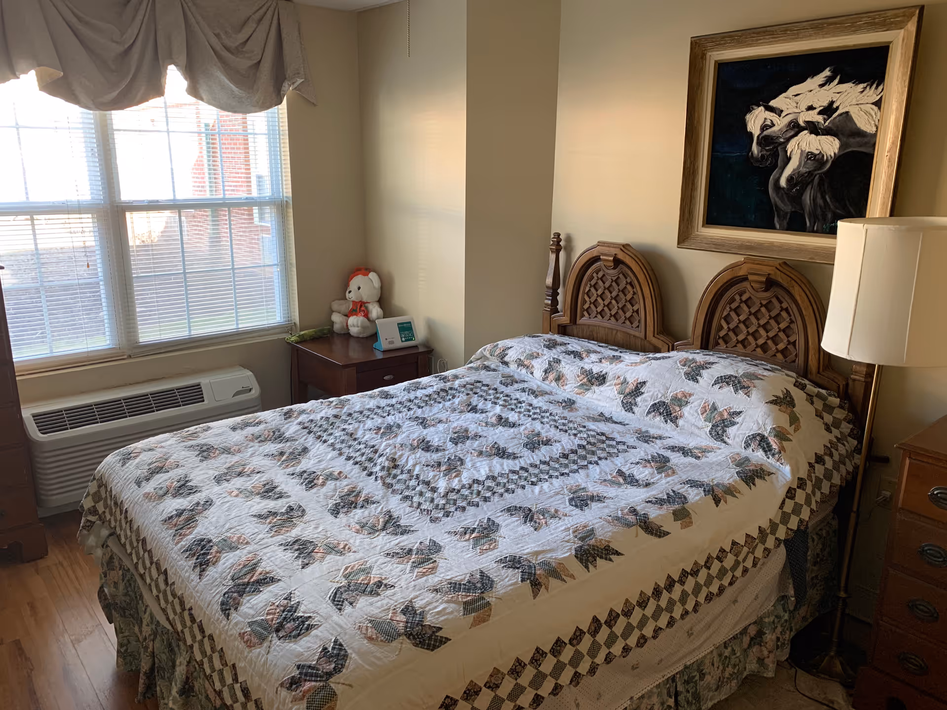 A bedroom with a wooden bed featuring a patterned quilt. There is a nightstand with a lamp on the right side of the bed and another nightstand with a teddy bear and a small device on the left side near a large window with blinds and a valance. A framed painting of three white horses hangs on the wall above the bed.
