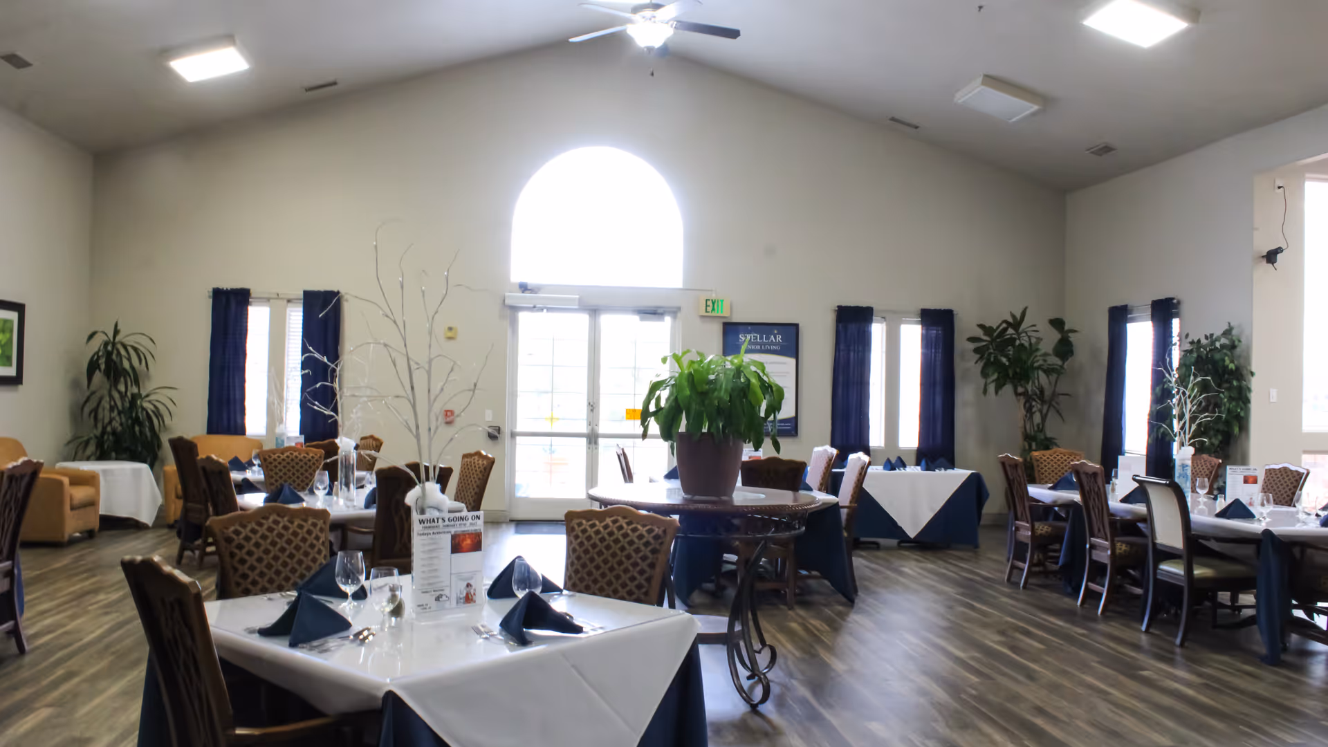 A spacious dining room with multiple tables covered in white and blue tablecloths, each set with folded navy napkins, glasses, and silverware. The room has large windows with dark curtains, potted plants, and a high ceiling with a ceiling fan. A round table in the center holds a large potted plant. The back wall features a large arched window and double glass doors with an exit sign above.