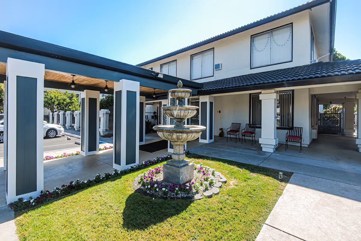 Outdoor view of Villa Serena Retirement Community featuring a three-tiered stone fountain surrounded by flowers and grass. The building has white walls with dark roof tiles and several windows. There is a covered walkway supported by white and dark blue pillars, with chairs placed near the entrance.