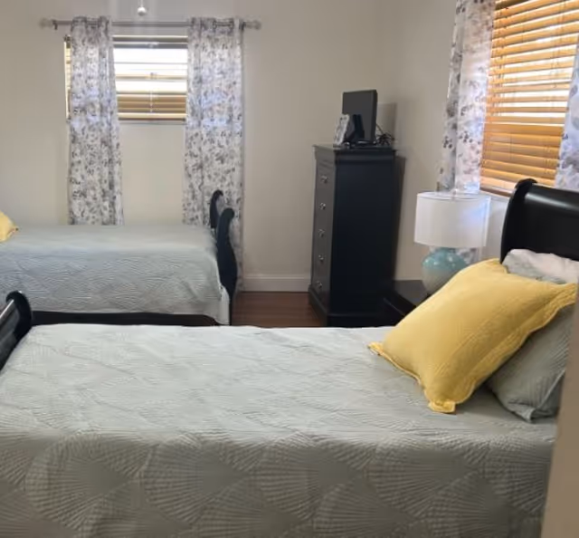 A bright bedroom with two twin beds dressed in light blue quilts and yellow pillows, a dresser, lamp, and windows with floral curtains.