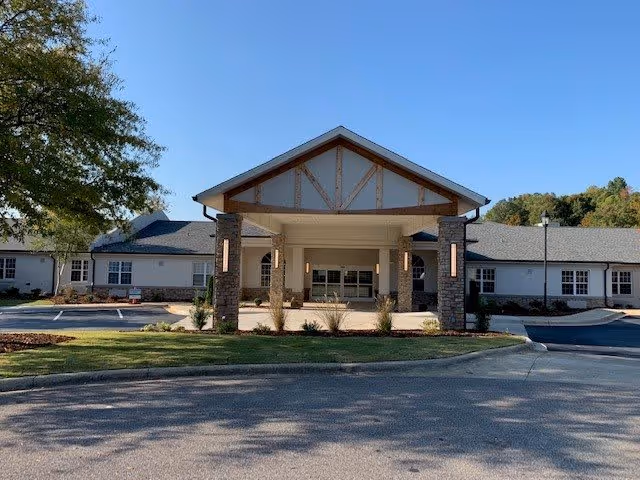 Front exterior view of The Ridge at Grandview facility showing a single-story building with a covered entrance supported by stone pillars, surrounded by a parking area and greenery under a clear blue sky.