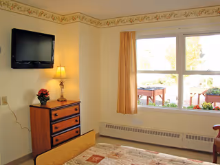 A small bedroom with a bed in the foreground, a wooden dresser with a lamp and a small plant on top, a wall-mounted flat-screen TV, and a large window with beige curtains letting in natural light. Outside the window, a garden area with plants and outdoor furniture is visible.