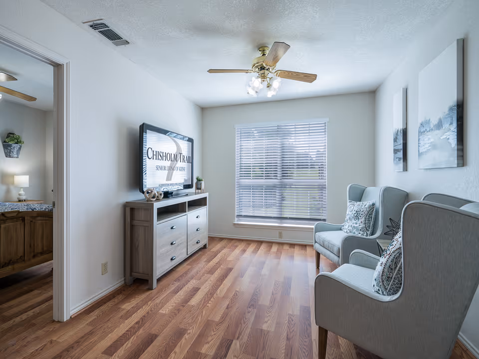 A bright living room with two light blue armchairs with patterned cushions facing a wall-mounted flat screen TV displaying 'Chisholm Trail Senior Living of Abilene'. The room has wooden flooring, a ceiling fan with lights, a large window with blinds, and two framed paintings on the wall. A doorway on the left leads to a bedroom with a wooden bed and a nightstand with a lamp.