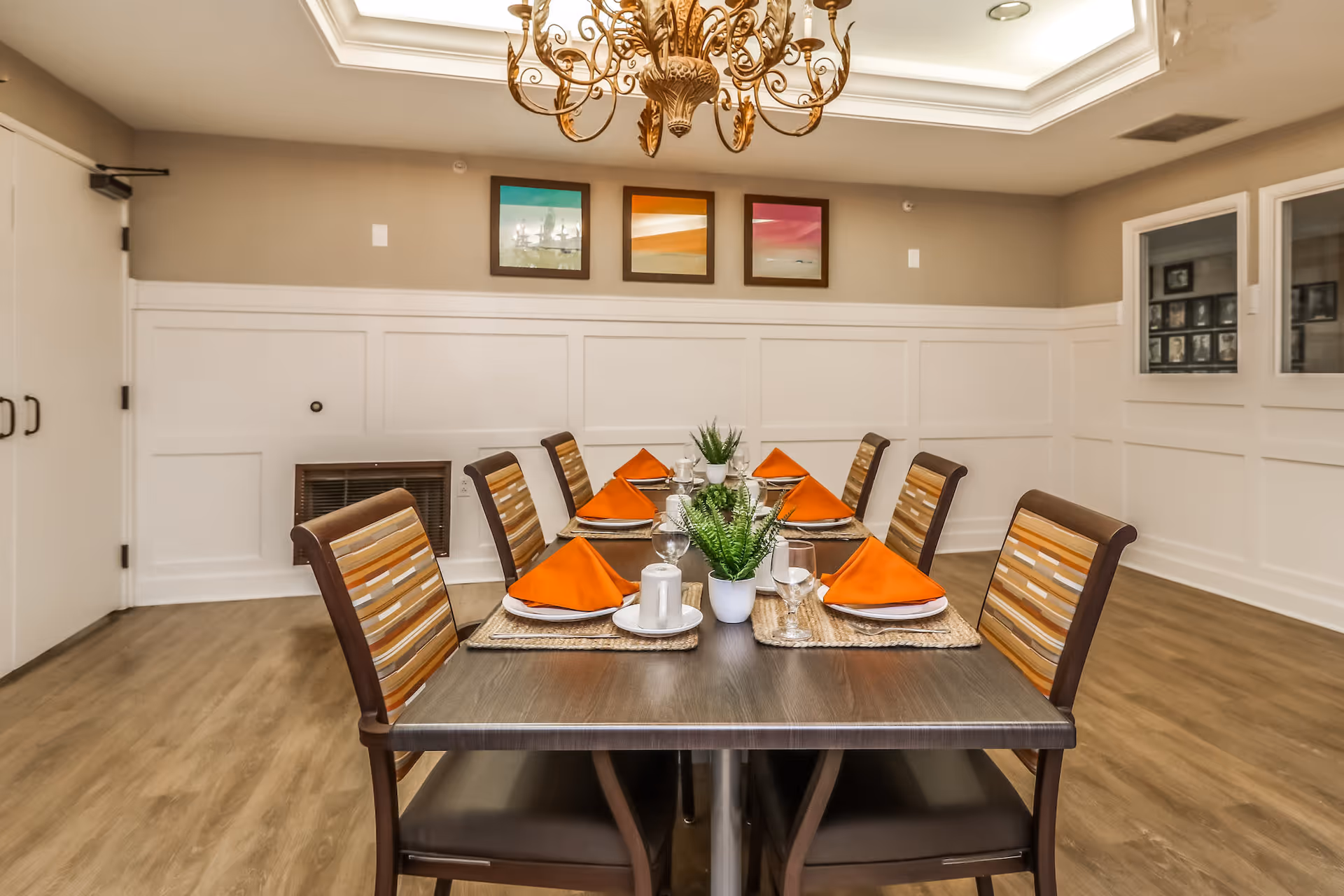 A dining room with a rectangular table set for six people. The table has orange folded napkins, plates, glasses, and small potted plants as centerpieces. The chairs have striped cushions. The room features wood flooring, white wainscoting on the walls, three framed pictures, and a decorative chandelier hanging from a tray ceiling.
