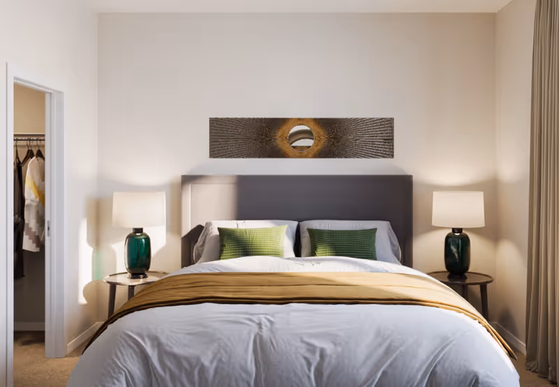 Well-lit bedroom with a made bed, green accent pillows, bedside lamps, wall art above the headboard, and an open closet doorway.