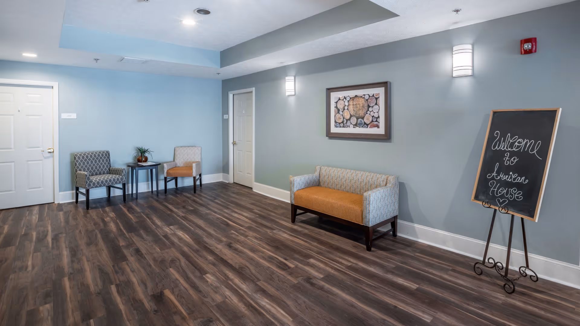 A waiting area with two patterned chairs and a small round table on the left, a two-tone upholstered bench on the right, a framed artwork on the wall, and a chalkboard sign that reads 'Welcome to American House' on a stand. The room has wood-look flooring and light blue walls with white trim.