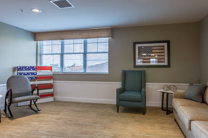 A small sitting room with armchairs, a sofa, framed American flag art, and a window with blinds.
