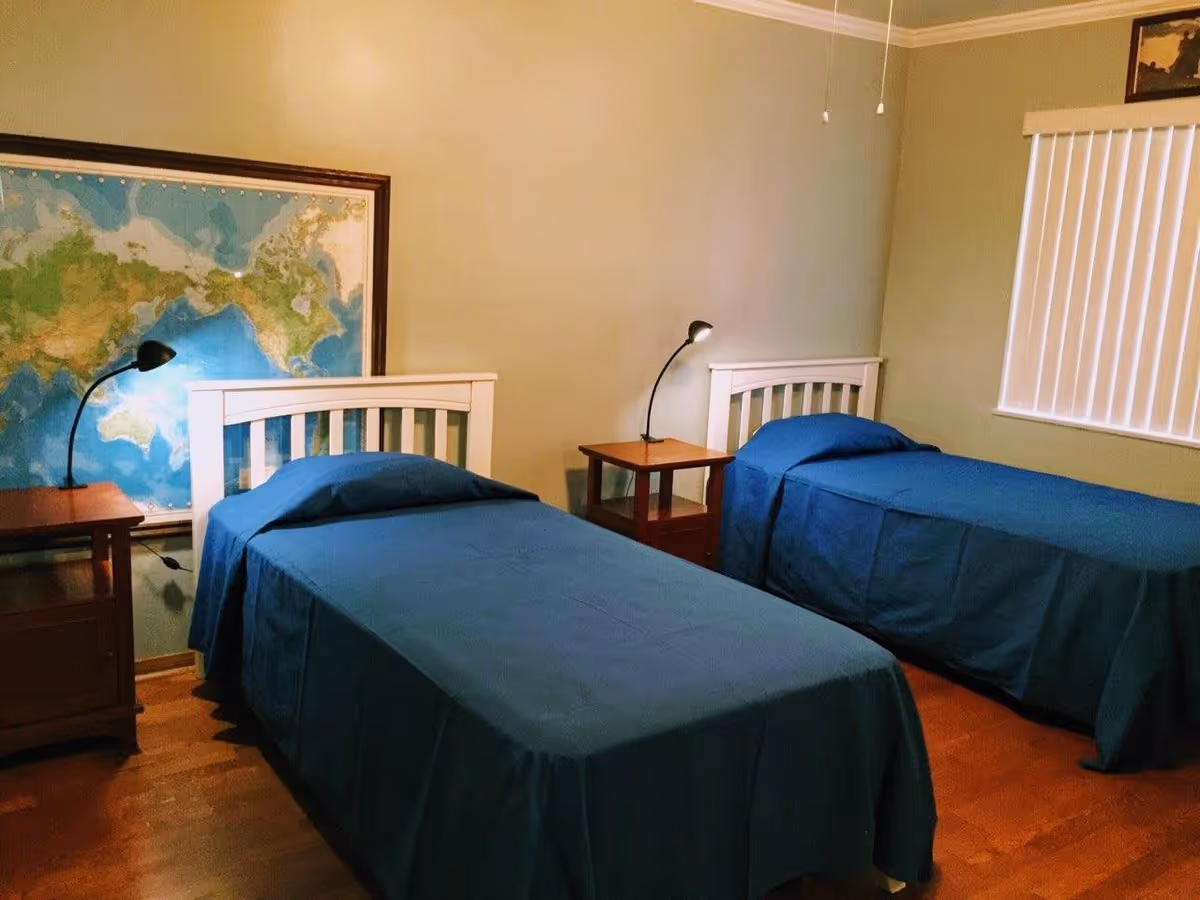 Two twin beds with blue bedding, nightstands and lamps in a simple bedroom with a world map on the wall.