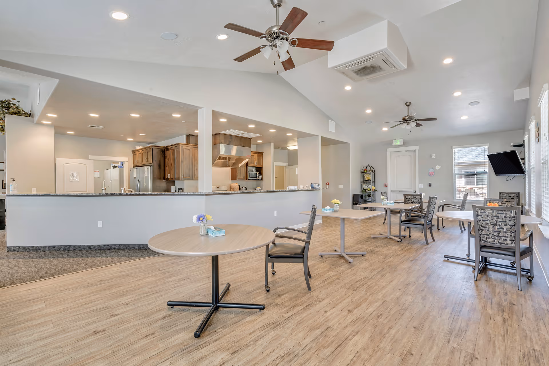 A spacious dining area with several round tables and chairs, each table decorated with a small flower vase and a tissue box. The room has wood flooring, ceiling fans, and recessed lighting. In the background, there is an open kitchen with wooden cabinets, stainless steel appliances, and a granite countertop that separates the kitchen from the dining area. Large windows allow natural light to fill the room.