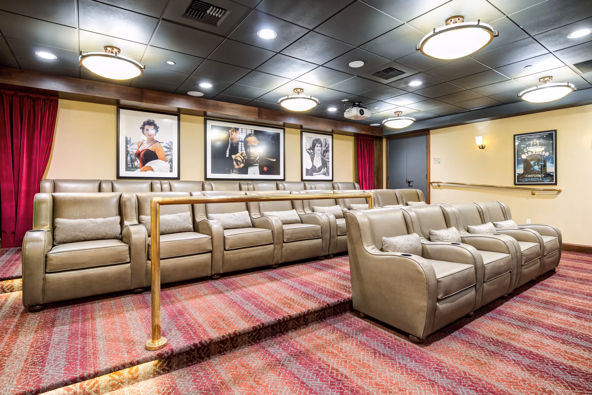A cozy home theater room with two rows of beige leather recliner chairs, each with a small pillow. The room has a red patterned carpet, a brass handrail separating the rows, and framed classic movie posters on the walls. The ceiling features recessed lighting and round ceiling lights.