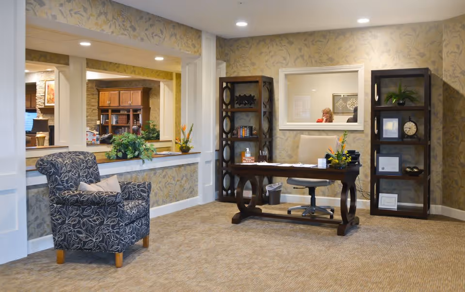 Reception area with a wooden desk and office chair, two dark wooden bookshelves with decorative items and plants, a patterned armchair with a pillow, and a window behind the desk showing a person inside. The walls have floral wallpaper and the floor is carpeted.
