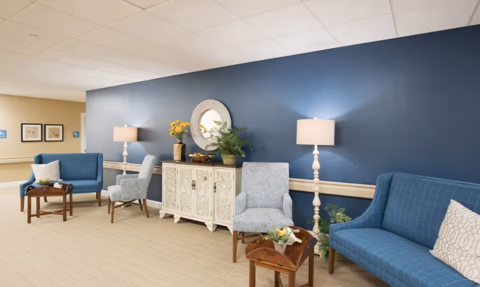 A cozy seating area in a senior living facility hallway with blue and patterned armchairs, blue sofas, wooden side tables with flower arrangements, two floor lamps, a white decorative cabinet with a round mirror above it, and a blue accent wall.