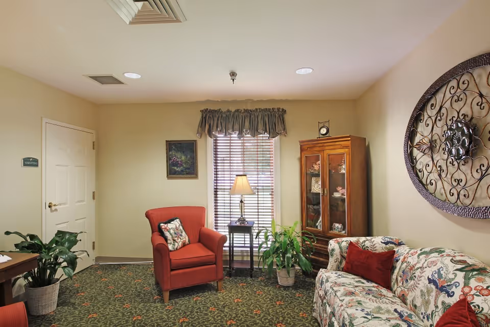 A cozy interior room with a red armchair, a floral patterned sofa with red cushions, a wooden cabinet with glass doors displaying decorative items, a small side table with a lamp, a potted plant, a window with blinds and a valance, and a decorative metal wall hanging. The room has beige walls and a patterned carpet.