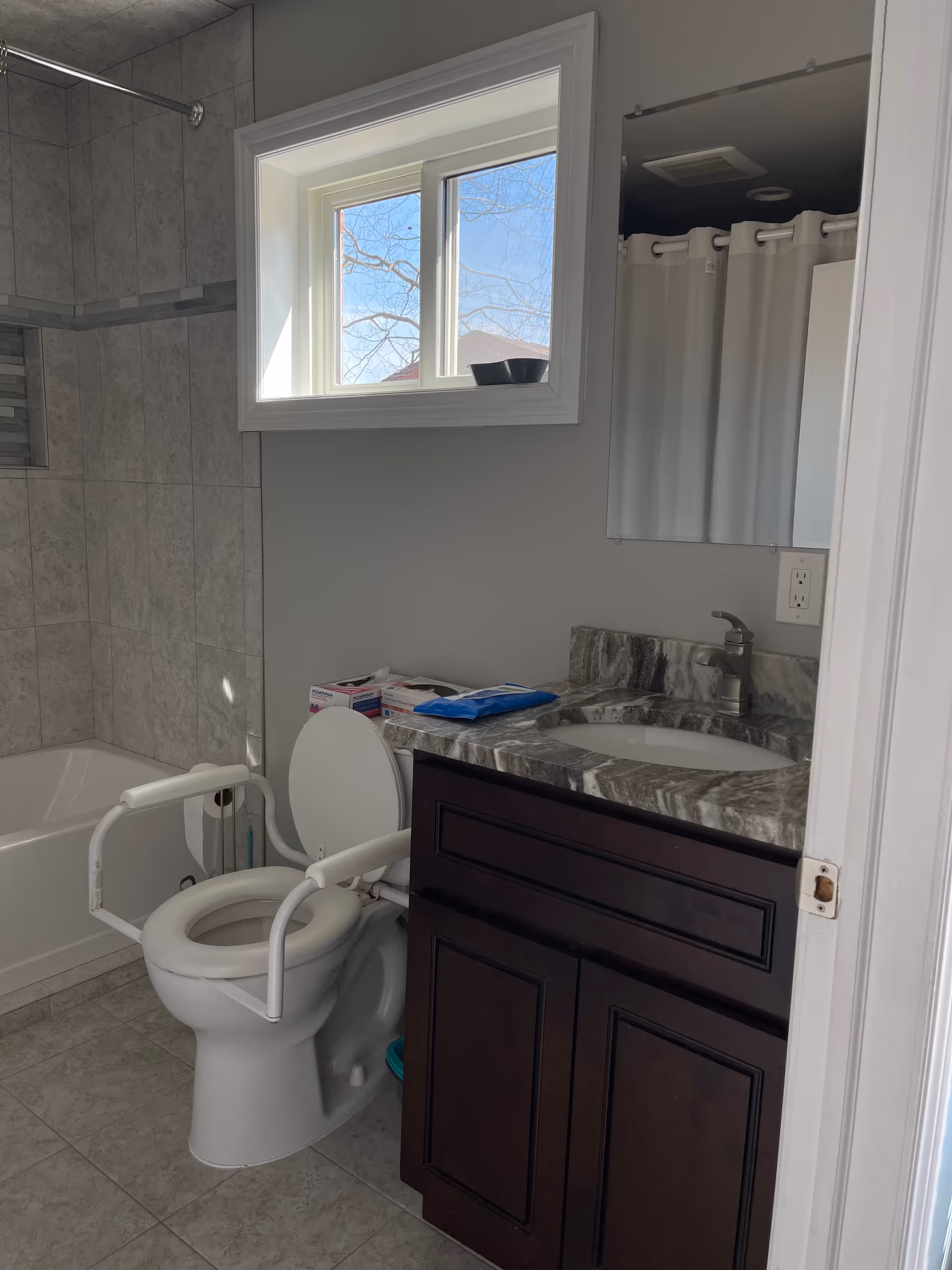 A bathroom with a toilet equipped with white safety rails, a bathtub with tiled walls, a window letting in natural light, and a dark wood vanity with a marble countertop and a sink. There are some hygiene products on the countertop and a mirror above the sink.