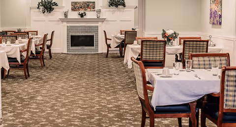 A dining room with multiple tables covered in white tablecloths, set with glasses, cups, and silverware. The chairs have wooden frames with plaid upholstery. In the background, there is a fireplace with decorative plants and a floral arrangement on the mantel. The room has a patterned carpet and light-colored walls with wainscoting.