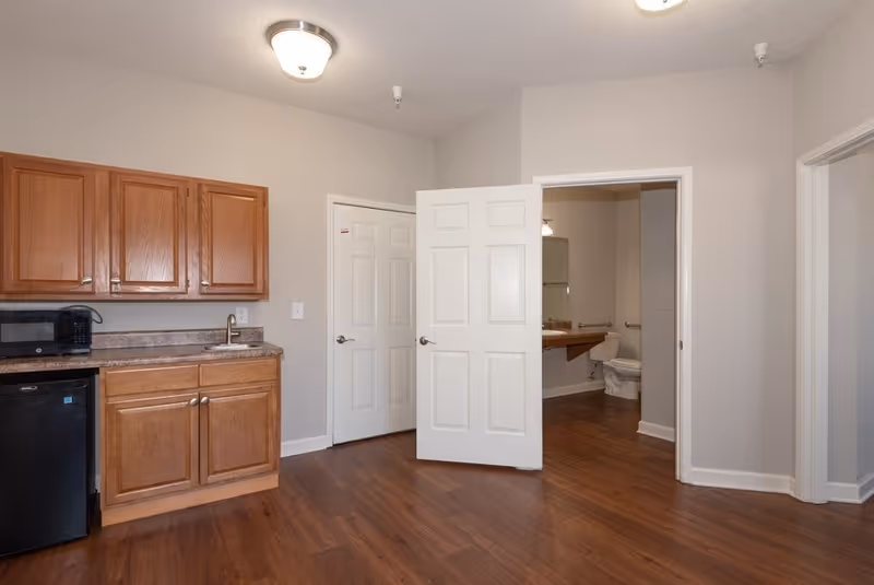 Small studio-style interior showing a kitchenette with wooden cabinets, microwave, mini-fridge, and an open door revealing a bathroom.