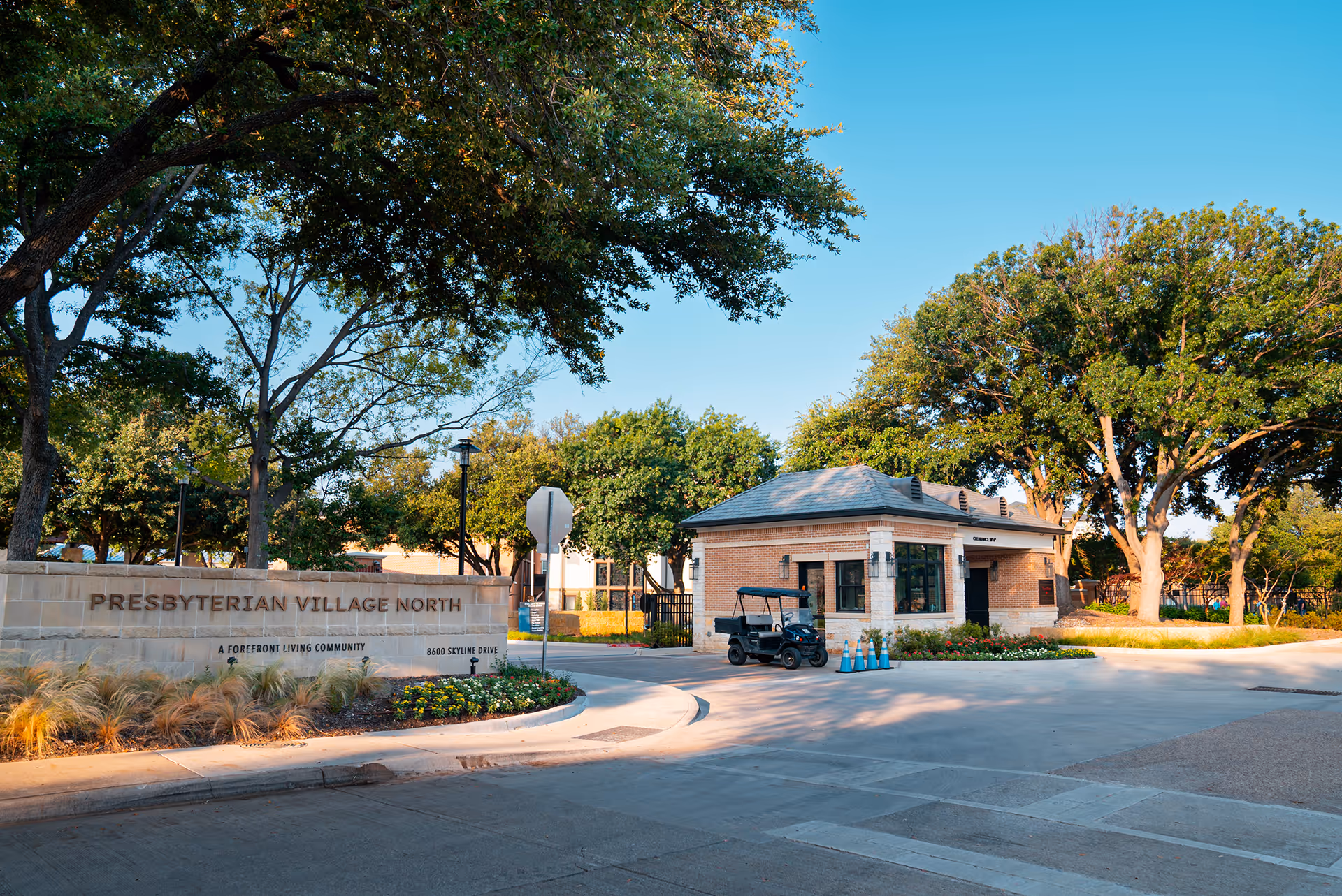 Entrance to Presbyterian Village North featuring a stone sign with the community name and a small brick guardhouse surrounded by trees and landscaping under a clear blue sky.