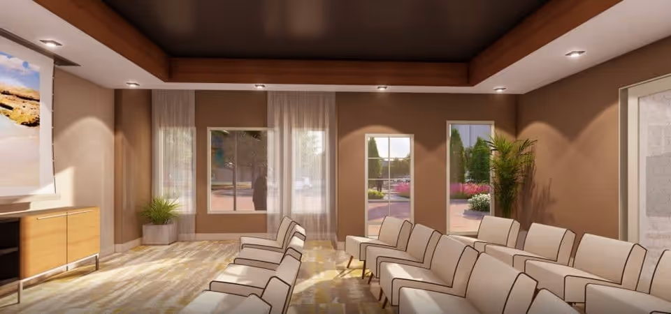 A well-lit waiting or lounge area with two rows of beige cushioned chairs facing a large screen on the left wall. The room has large windows and a glass door letting in natural light, with plants placed near the windows and door. The ceiling features recessed lighting and a wooden tray design.