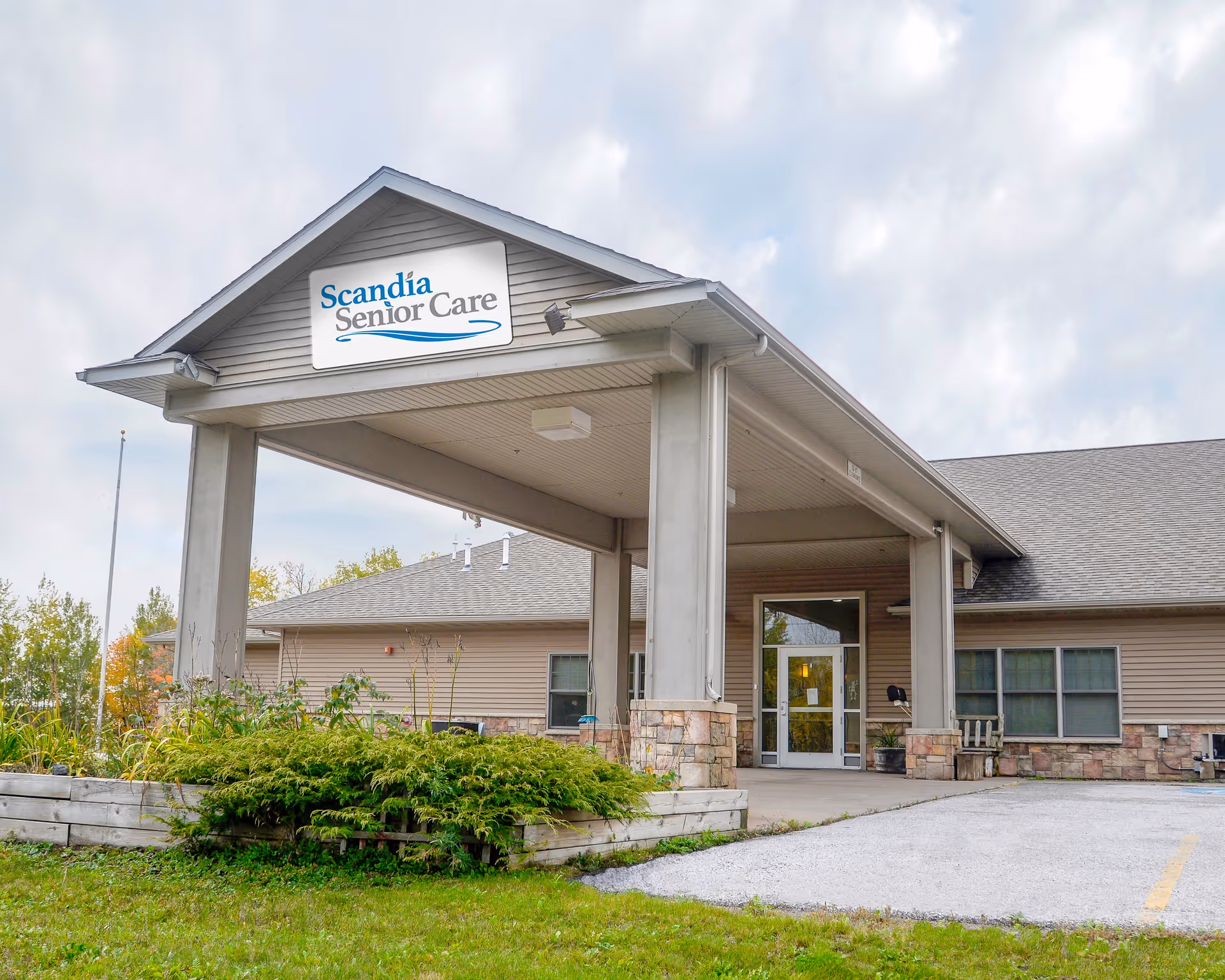 Exterior view of a senior care facility building with a covered entrance. The building has beige siding, stone accents at the base, and a sign above the entrance that reads 'Scandia Senior Care'. There is greenery and a small garden bed in front of the entrance, with a cloudy sky in the background.