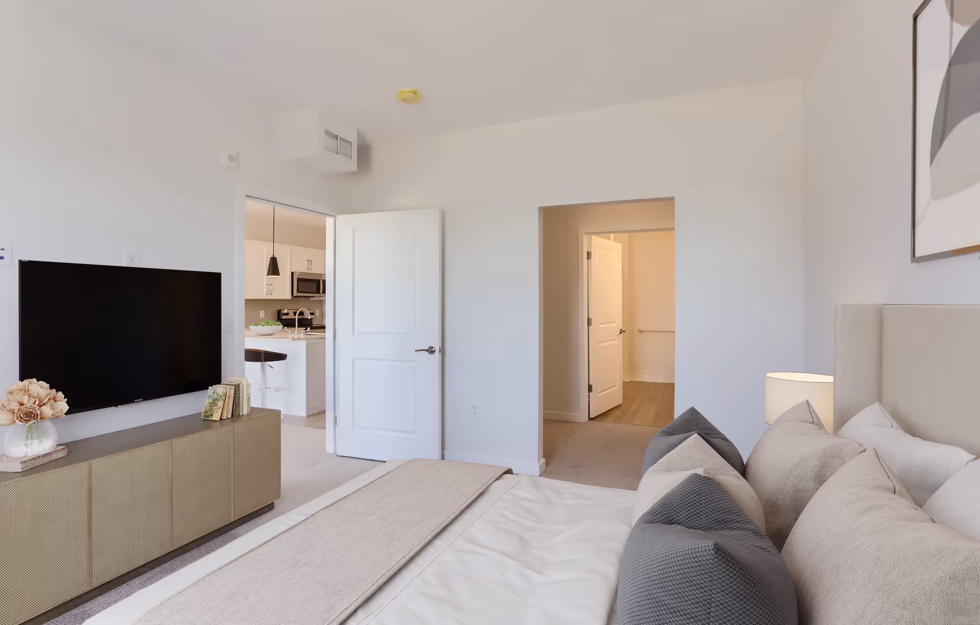 A modern bedroom with a beige upholstered bed adorned with multiple pillows and a beige throw blanket. A wooden cabinet with a vase of flowers and books sits beneath a mounted flat-screen TV. The room has white walls and two open doorways, one leading to a kitchen area with white cabinetry and a bar stool, and the other leading to a hallway with a closed door. A framed abstract artwork hangs on the wall above the bed.