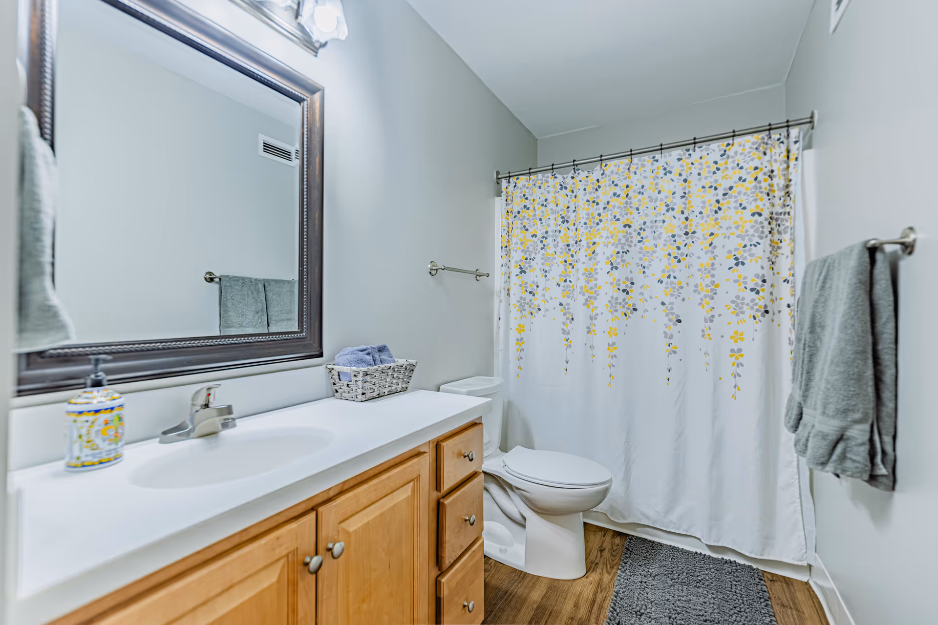 A clean bathroom with a wooden vanity featuring a white countertop and an oval sink. Above the vanity is a large framed mirror and a light fixture. To the right is a toilet next to a bathtub with a white shower curtain decorated with yellow and gray floral patterns. Gray towels hang on towel racks, and a gray bath mat is on the wooden floor.