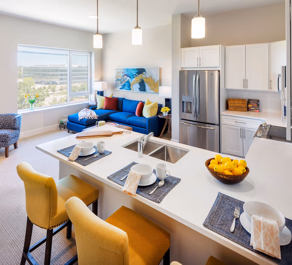 Bright open-concept living area with a blue sofa by a large window and a kitchen island set with place settings and yellow bar stools.
