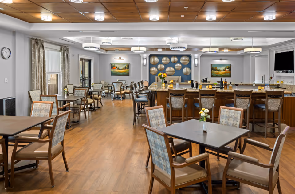 A spacious dining area in a senior living facility featuring multiple tables with chairs, a large bar counter with high stools, decorative wall art, and ceiling lights. The room has wooden flooring, light-colored walls, and windows with curtains allowing natural light.
