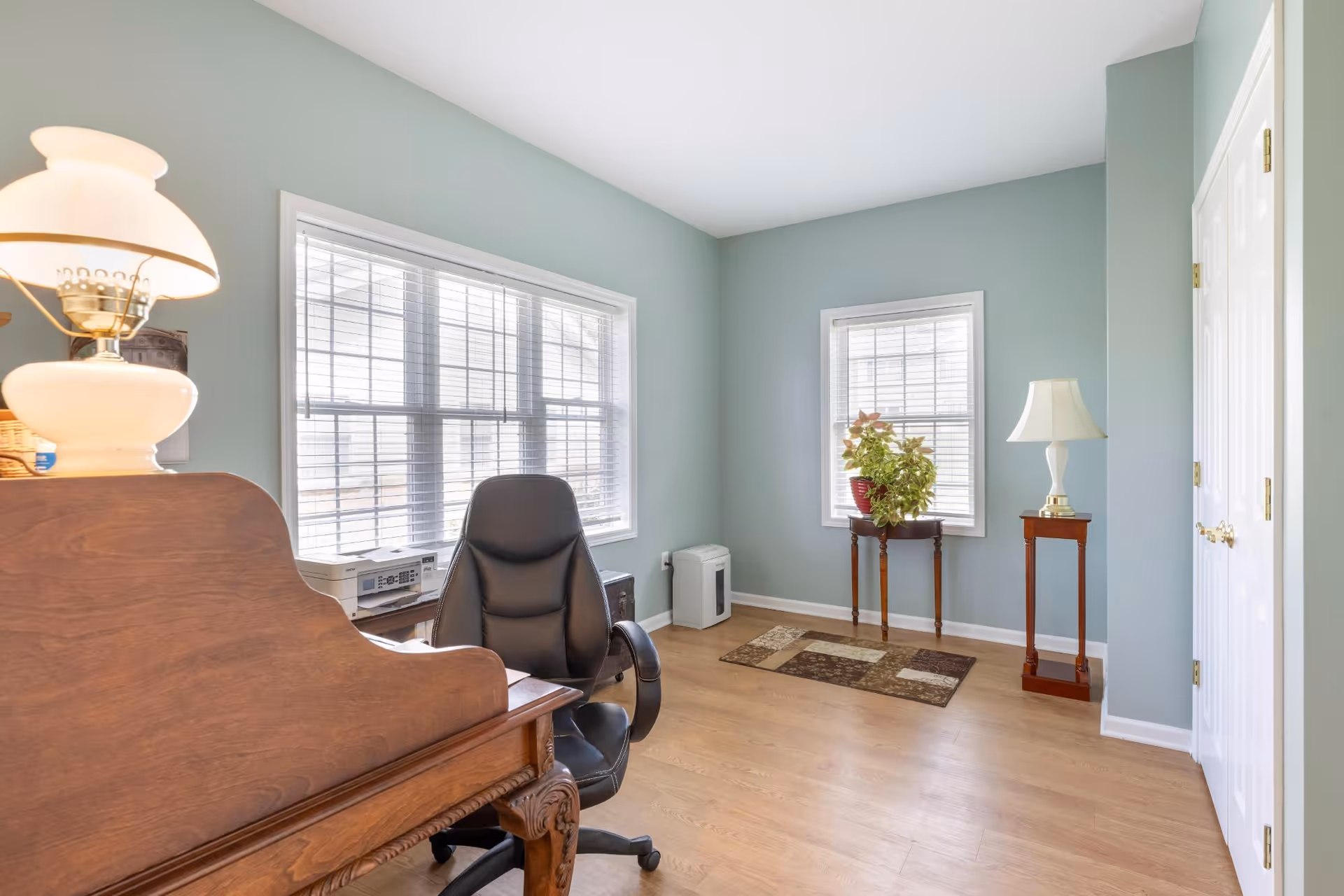 A bright room with light blue walls and wooden flooring featuring a wooden desk with a lamp and printer on it, a black office chair, two windows with white blinds, a small table with a potted plant, a floor lamp on a wooden stand, and a patterned rug on the floor.