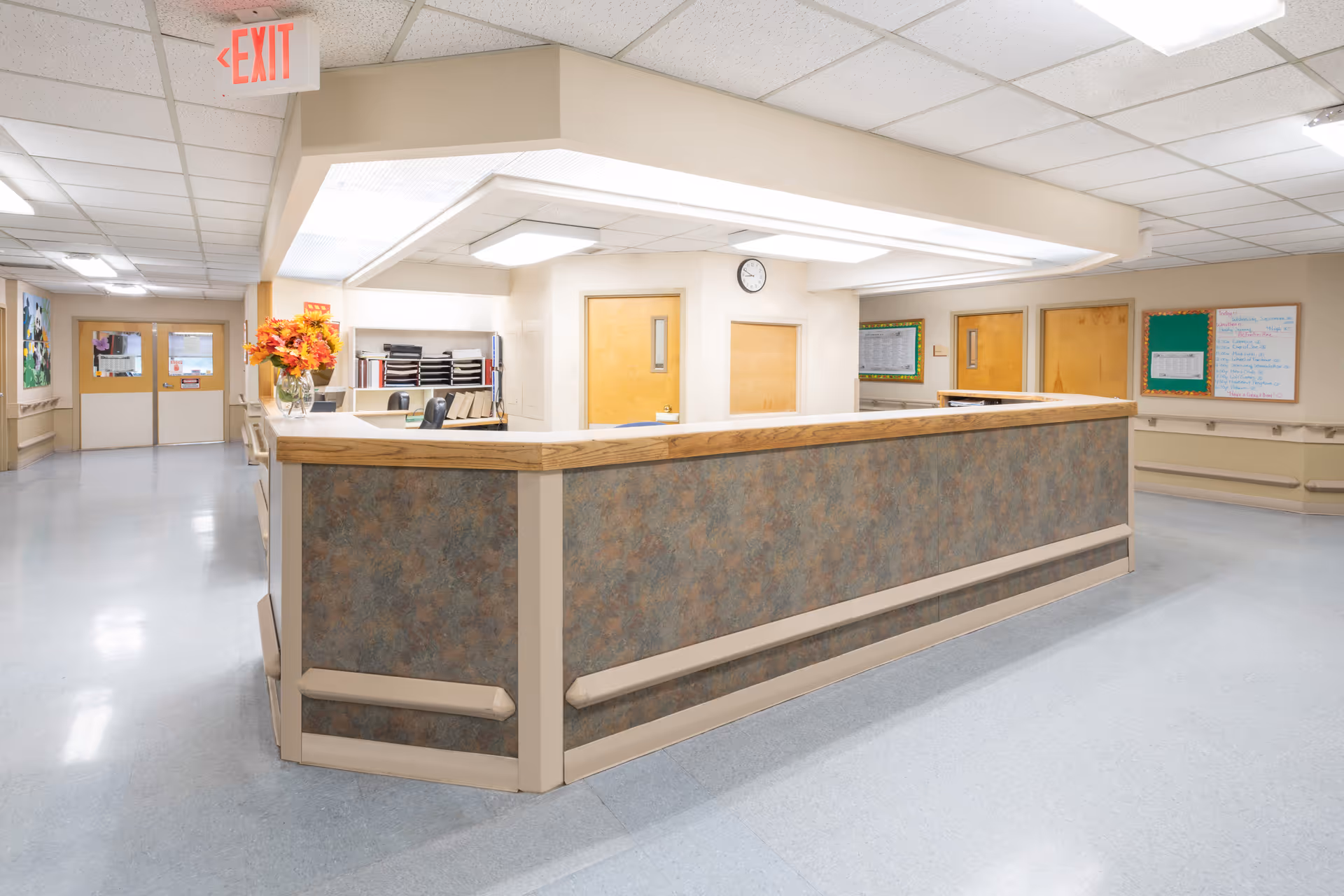 A clean and well-lit nursing station area inside Dresher Hill Health & Rehabilitation Center. The station has a large counter with a wood trim and a textured panel front. Behind the counter are office supplies, shelves with files, and a clock on the wall. The surrounding hallway has beige walls, multiple doors, bulletin boards, and an illuminated exit sign overhead.