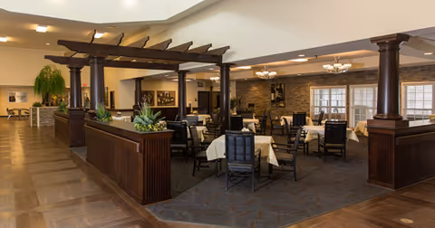 A spacious dining area with multiple tables covered with white tablecloths and surrounded by dark wooden chairs. The room features dark wooden columns and a pergola-style divider with plants on top. The floor is a combination of wood and carpet, and the walls have large windows and stone accents. Ceiling lights provide warm illumination.