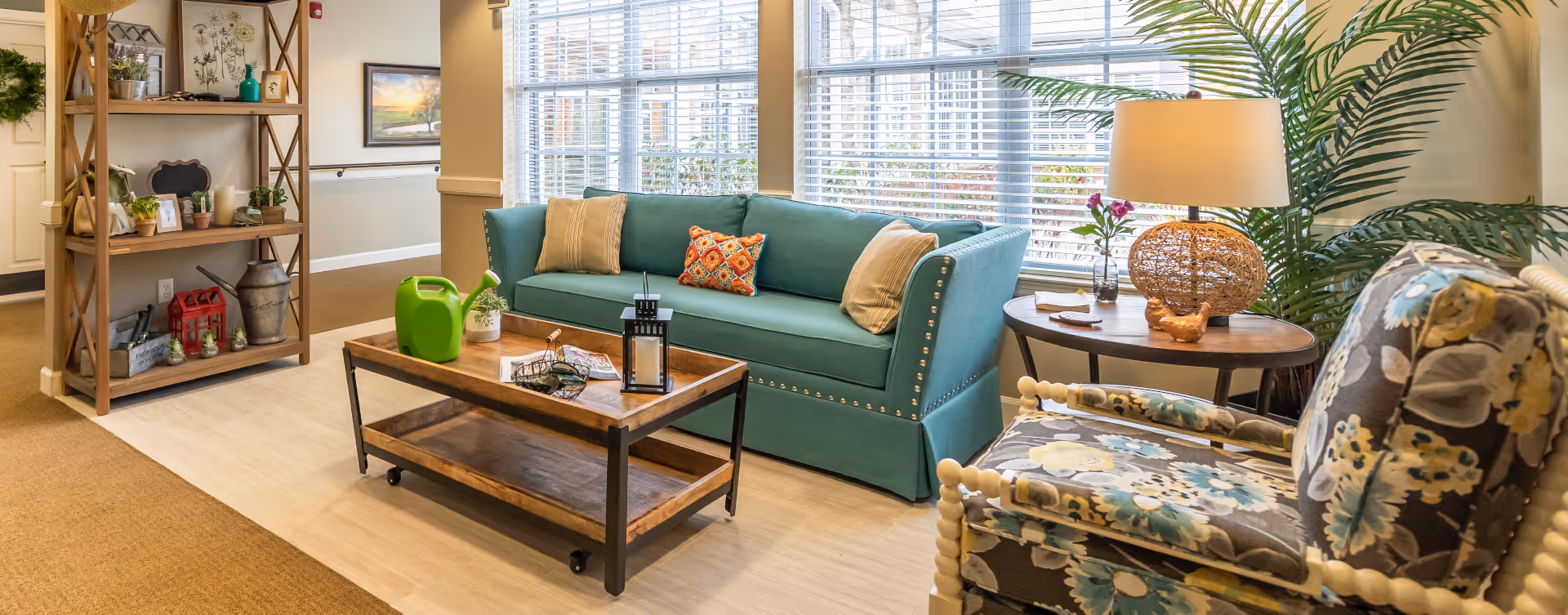 A cozy living room area with a teal sofa adorned with three decorative pillows, a wooden coffee table with a green watering can and a lantern, a floral patterned armchair, a round side table with a lamp and a small plant, and a wooden shelving unit with various decorative items. Large windows with blinds allow natural light to fill the space.