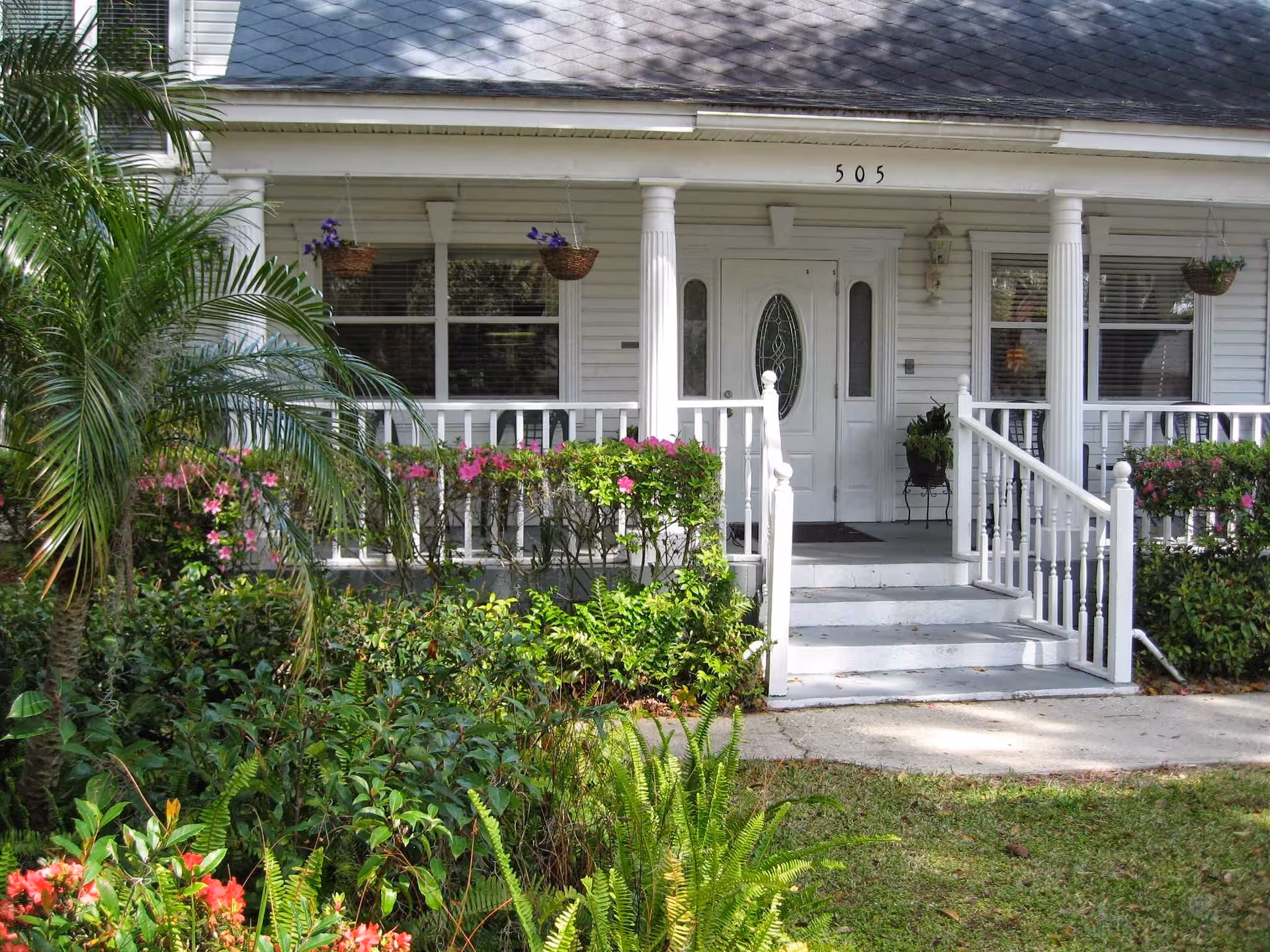 Front exterior view of a white house with a porch featuring white railings and columns. There are hanging flower baskets and bushes with pink flowers in front of the porch. The house number 505 is visible above the door.