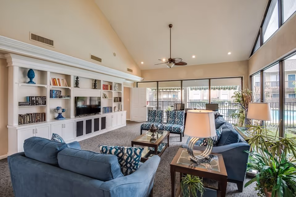 A spacious living room with high vaulted ceilings, large windows letting in natural light, blue sofas, patterned chairs, a glass coffee table, side tables with lamps, built-in white shelves with books and decorative items, and a ceiling fan. Outside the windows, a swimming pool and apartment buildings are visible.