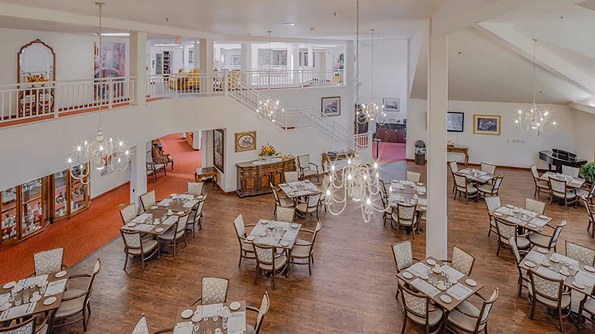 Spacious dining room in a senior living facility with many set tables, chandeliers, wood floors, and an upper balcony.