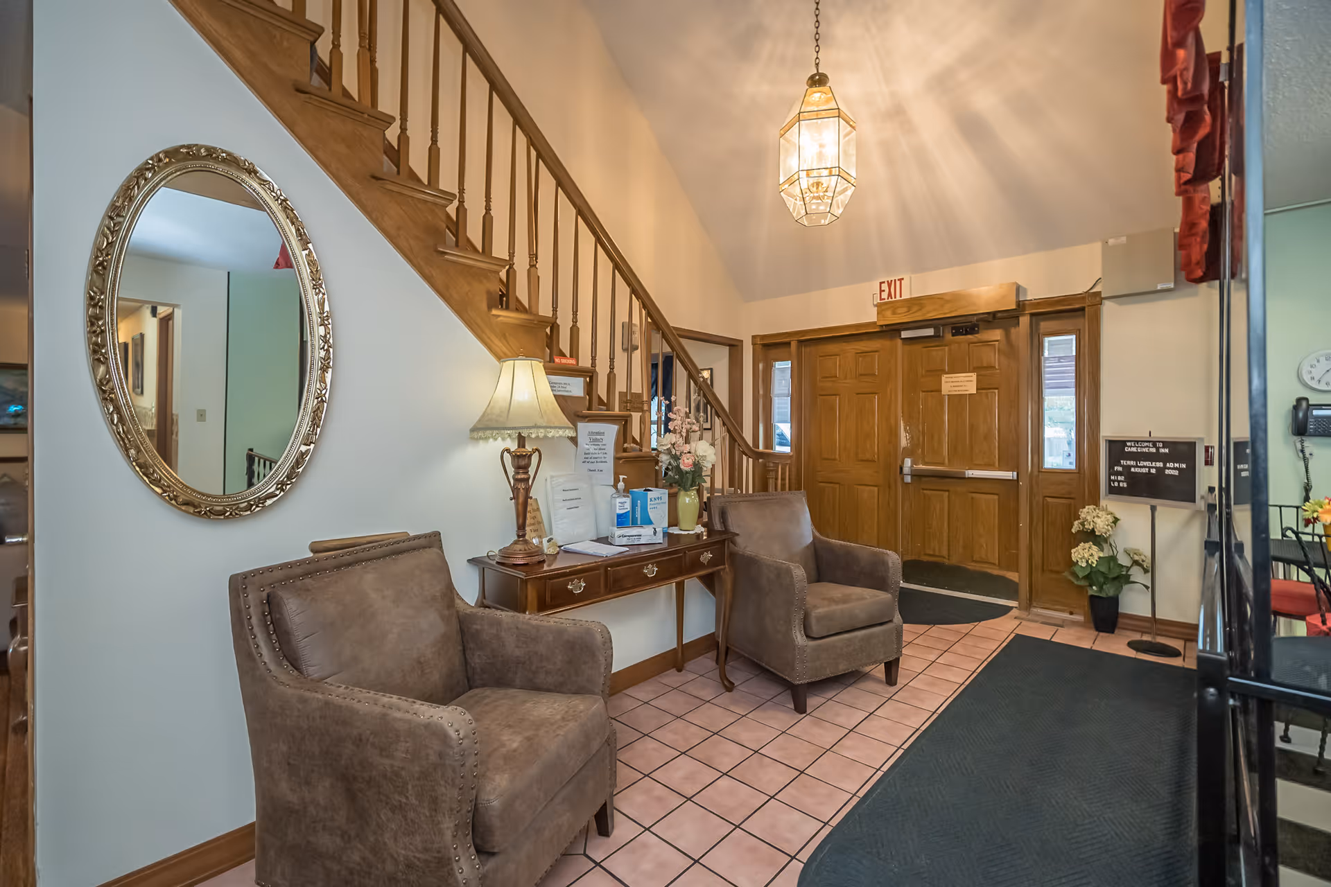 An interior view of a facility entrance area with two brown upholstered armchairs, a wooden side table with a lamp and flowers, a large oval mirror on the wall, a staircase with wooden railing, and double wooden exit doors. There is a hanging light fixture on the ceiling and a small signboard near the door.