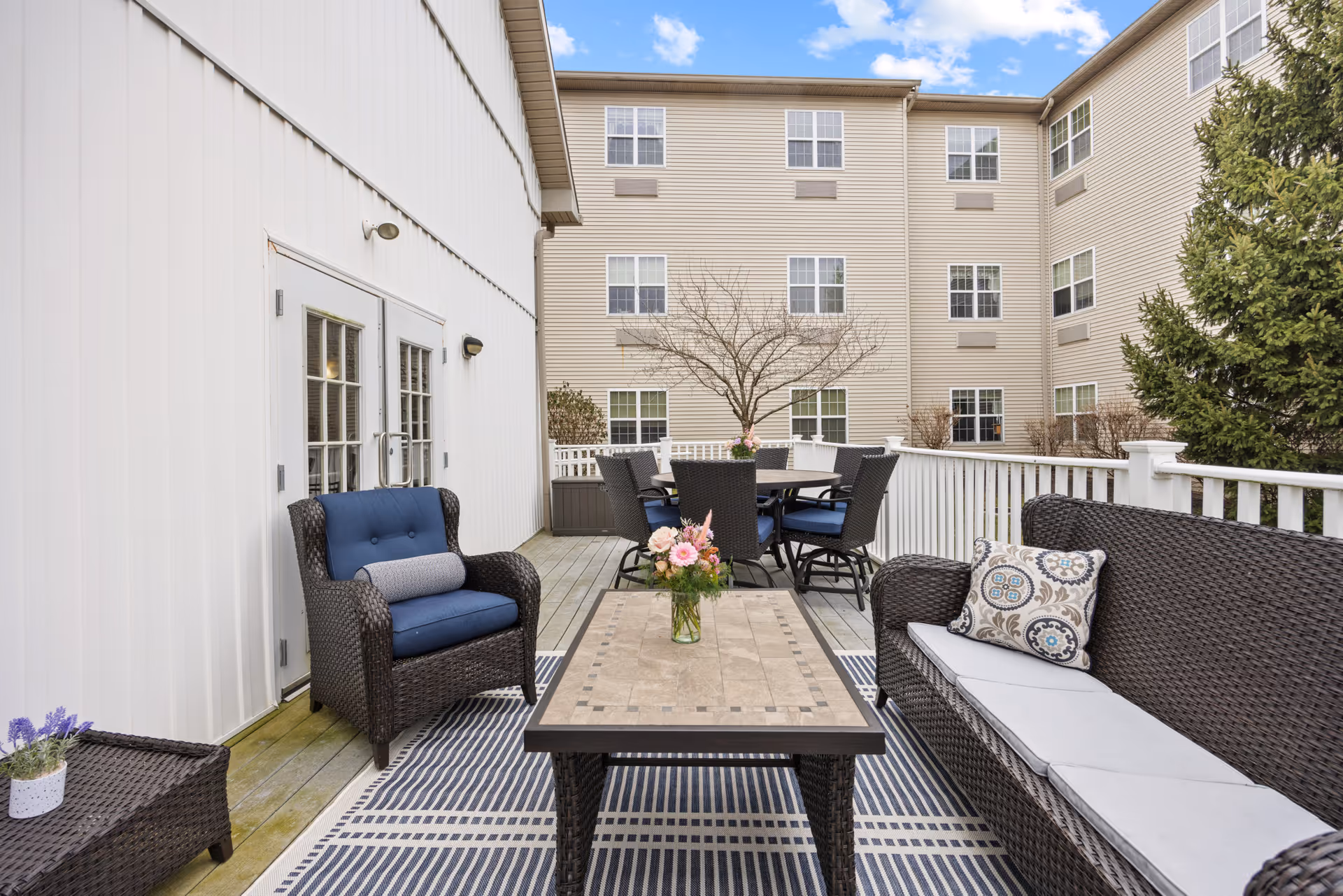 Outdoor patio area with wicker furniture including a cushioned armchair, a cushioned sofa with a decorative pillow, and a rectangular table with a flower vase on top. In the background, there is a round dining table with six chairs. The patio is surrounded by a white railing and adjacent to a beige multi-story building with multiple windows. A tree without leaves and some greenery are also visible.