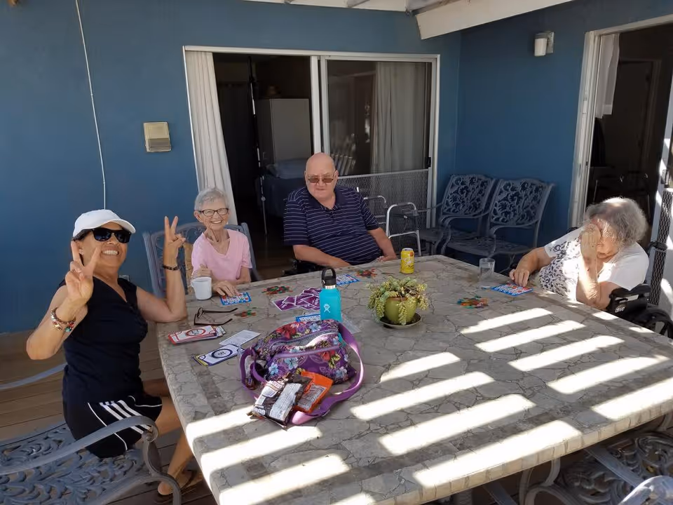 Four older adults seated around a sunlit outdoor stone patio table playing games and socializing.
