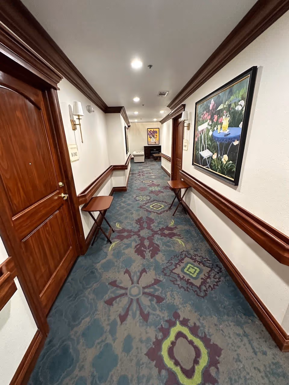 A well-lit indoor hallway with patterned carpet in shades of blue, purple, and green. The walls are white with dark wood trim and handrails. There are wooden doors on both sides, small folding tables attached to the walls, and framed paintings hanging on the walls. At the end of the hallway, there is a cushioned bench and a cabinet with a floral painting above it.