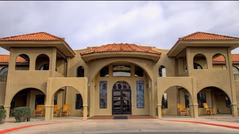 Front exterior view of The Garnet of Casa Grande building with a beige stucco facade, red tile roof, arched entryways, and rocking chairs on the porch.