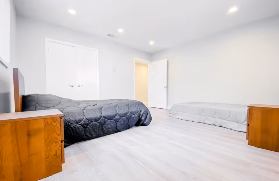 A bright, spacious bedroom with light-colored walls and flooring. The room contains two beds, one with a dark gray quilt and the other with a light gray quilt. Each bed has a wooden nightstand beside it. The room has recessed ceiling lights, a closed double-door closet, and an open door leading to another area.